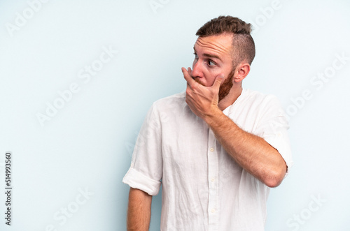 Young caucasian man isolated on blue background thoughtful looking to a copy space covering mouth with hand. © Asier