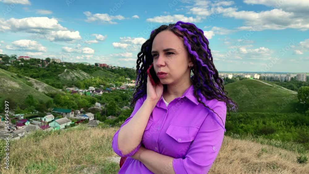 Young beautiful woman talking on mobile phone, standing in hilly terrain. Cute vivid female with dreadlocks speaking on smartphone, standing outdoors in cloudy, windy, summer weather. Young beautiful woman talking on mobile phone, standing in hilly terrain. Cute vivid female with dreadlocks speaking on smartphone, standing outdoors in cloudy, windy, summer weather.