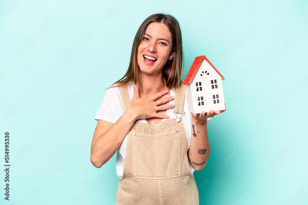Young caucasian pregnant woman holding an toy house isolated on blue background laughs out loudly keeping hand on chest. Young caucasian pregnant woman holding an toy house isolated on blue background laughs out loudly keeping hand on chest.
