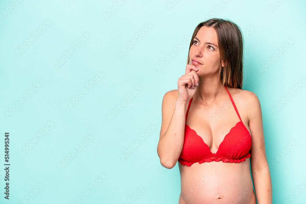 Fototapeta premium Young caucasian pregnant woman wearing bikini isolated on blue background looking sideways with doubtful and skeptical expression.