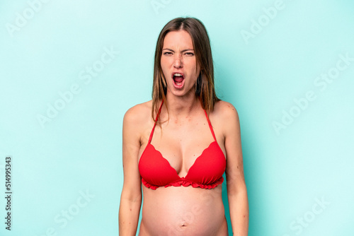 Young caucasian pregnant woman wearing bikini isolated on blue background screaming very angry and aggressive. © Asier