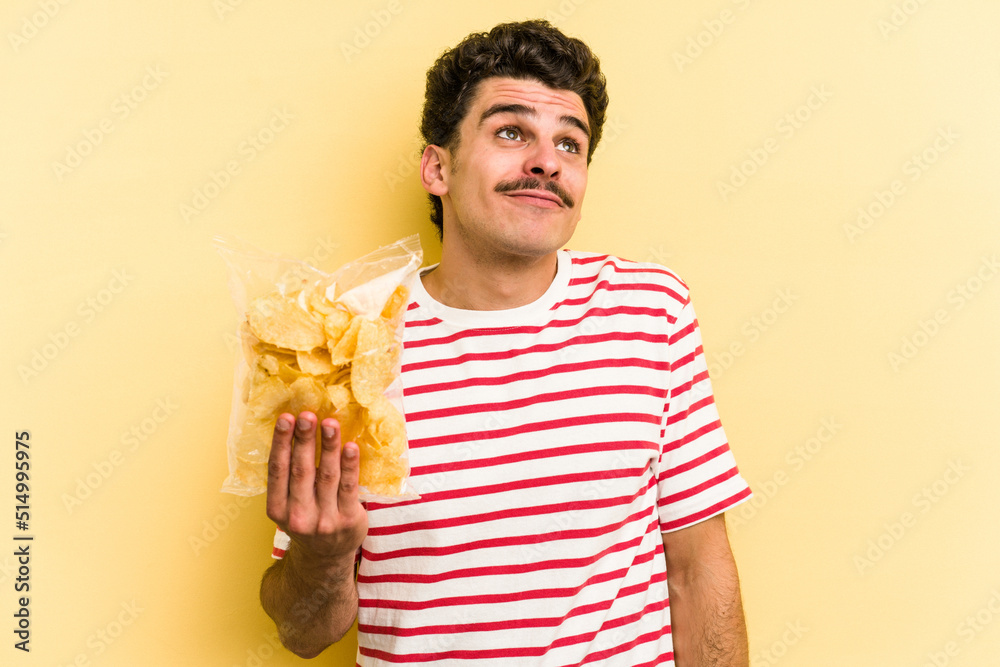 Young caucasian man holding a bag of chips isolated on yellow background dreaming of achieving goals and purposes Young caucasian man holding a bag of chips isolated on yellow background dreaming of achieving goals and purposes