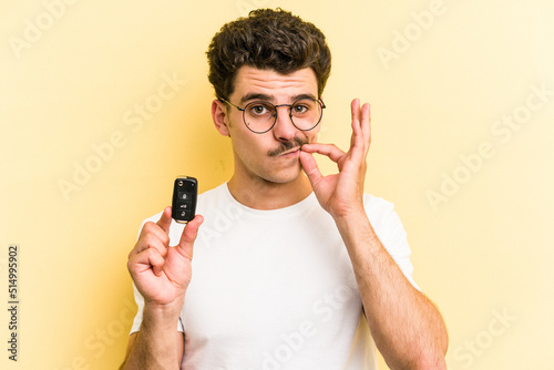 Young caucasian man holding car keys isolated on yellow background with fingers on lips keeping a secret. © Asier