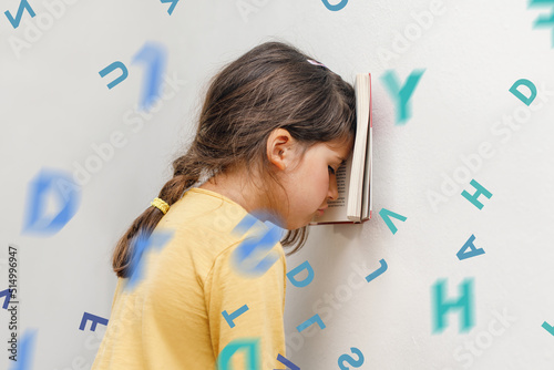 Sad and tired caucasian girl with dyslexia holds a book on her forehead. Flying tangled letters in the air. The child learns to speak and read correctly photo