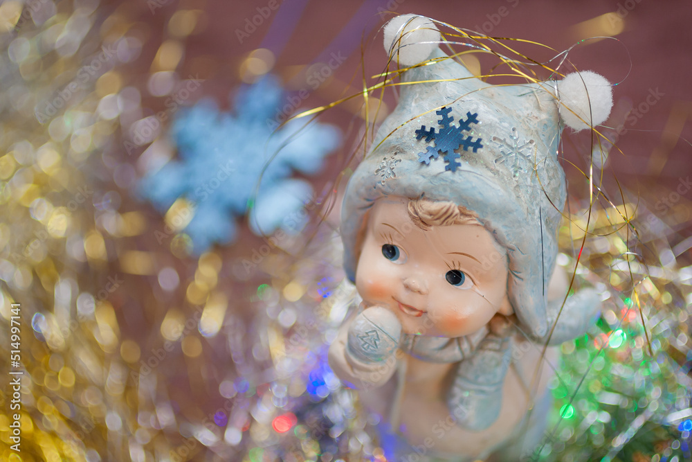 Top view of the ceramic figure of a cute little child in the winter clothes with the glittering snowflake and the golden tinsel on his cap. New year, Christmas background with the space for text Top view of the ceramic figure of a cute little child in the winter clothes with the glittering snowflake and the golden tinsel on his cap. New year, Christmas background with the space for text