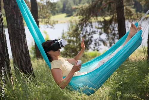 middle aged woman playing game in virtual reality glasses in a hammock relaxed outdoors. copy space.