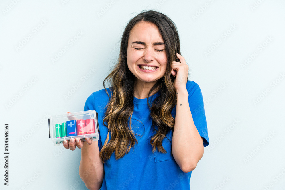 Young caucasian woman holding batteries isolated on blue background covering ears with hands. Young caucasian woman holding batteries isolated on blue background covering ears with hands.