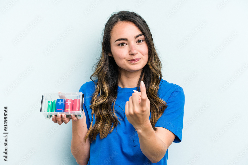 Young caucasian woman holding batteries isolated on blue background pointing with finger at you as if inviting come closer. Young caucasian woman holding batteries isolated on blue background pointing with finger at you as if inviting come closer.