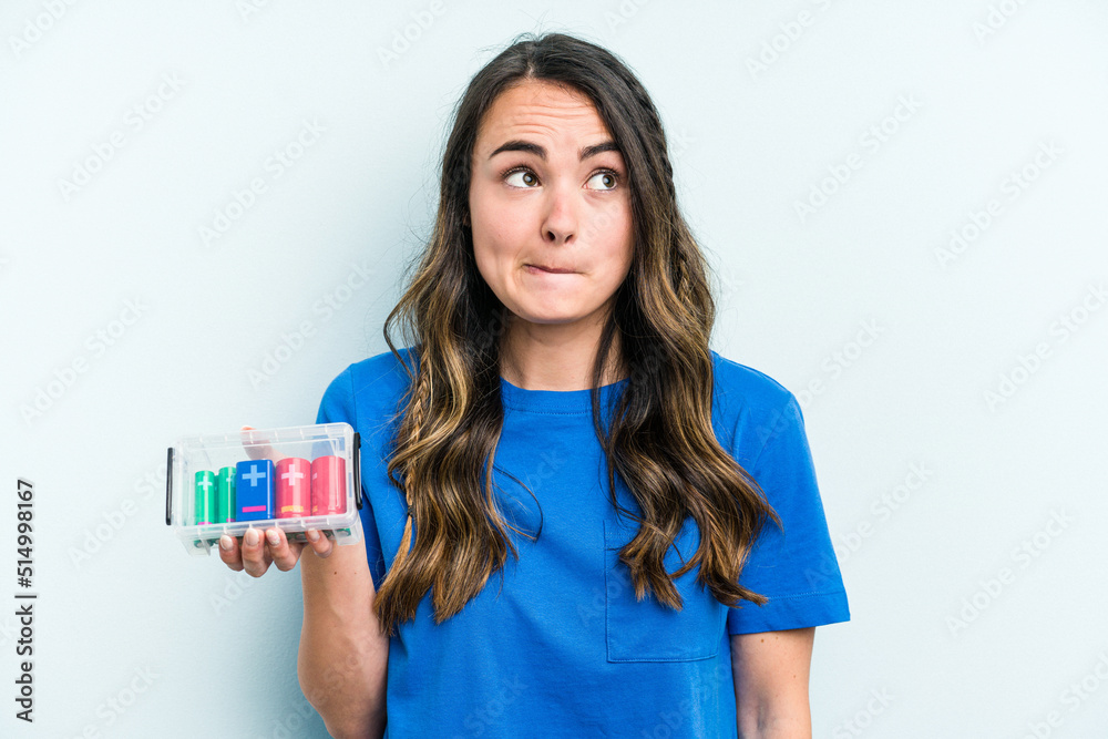 Young caucasian woman holding batteries isolated on blue background confused, feels doubtful and unsure. Young caucasian woman holding batteries isolated on blue background confused, feels doubtful and unsure.