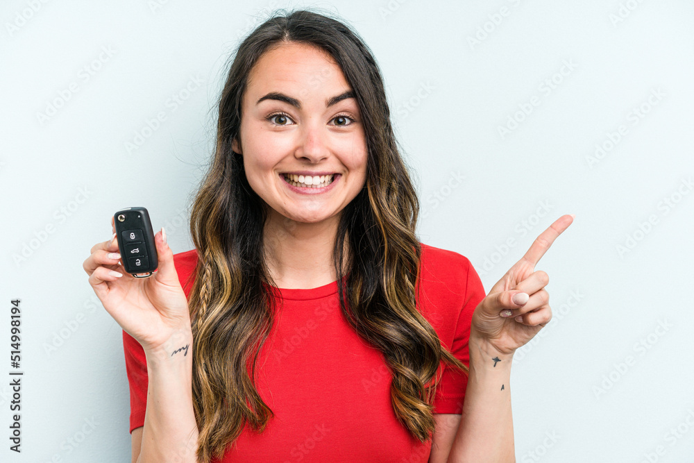 Young caucasian woman holding car keys isolated on blue background smiling and pointing aside, showing something at blank space. Young caucasian woman holding car keys isolated on blue background smiling and pointing aside, showing something at blank space.