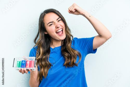 Young caucasian woman holding batteries isolated on blue background raising fist after a victory, winner concept.