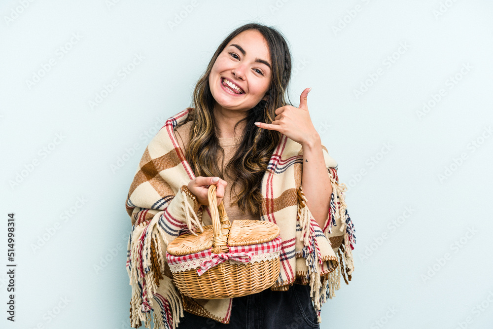 Young caucasian woman holding a picnic basket isolated on blue background showing a mobile phone call gesture with fingers. Young caucasian woman holding a picnic basket isolated on blue background showing a mobile phone call gesture with fingers.