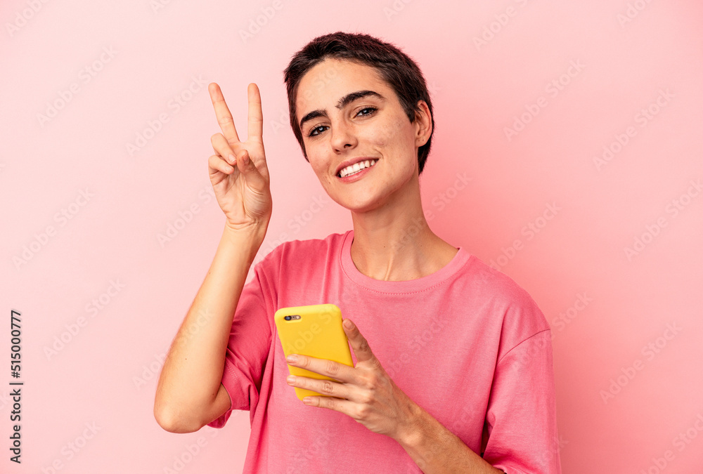 Young caucasian woman holding mobile phone isolated on pink background joyful and carefree showing a peace symbol with fingers. Young caucasian woman holding mobile phone isolated on pink background joyful and carefree showing a peace symbol with fingers.