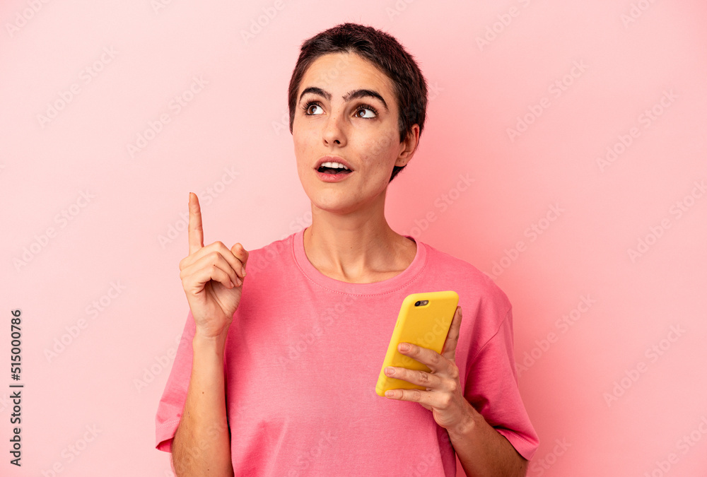 Young caucasian woman holding mobile phone isolated on pink background pointing upside with opened mouth. Young caucasian woman holding mobile phone isolated on pink background pointing upside with opened mouth.