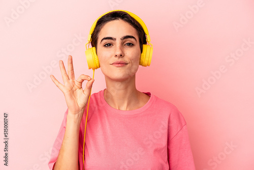 Young caucasian woman listening to music isolated on pink background cheerful and confident showing ok gesture.