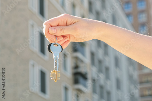 Woman's hand holds a key against the backdrop of a multi-storey residential area. The concept of buying residential real estate, winning the lottery, affordable mortgage
