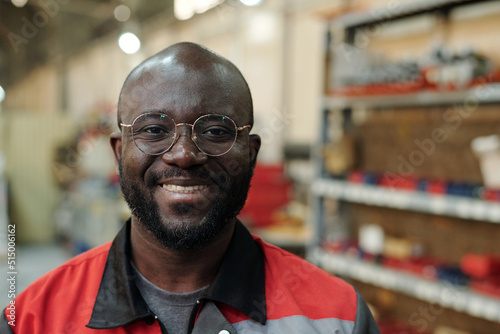 Young successful black man in uniform and eyeglasses looking at camera while standing against shelves with working materials