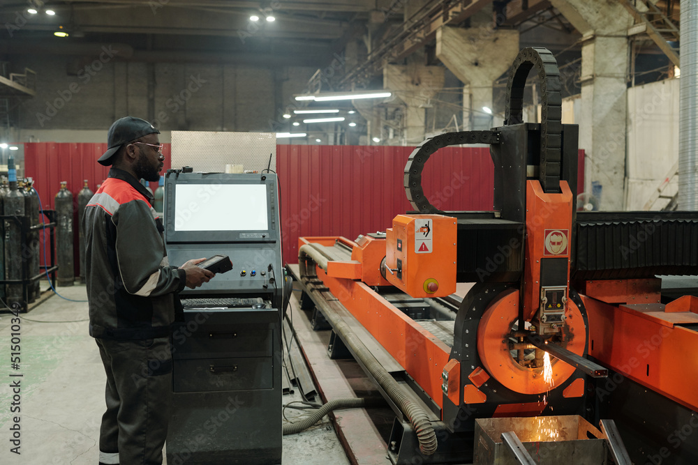 Fototapeta premium Young male worker of industrial plant standing by control terminal while checking work of production machine in workshop