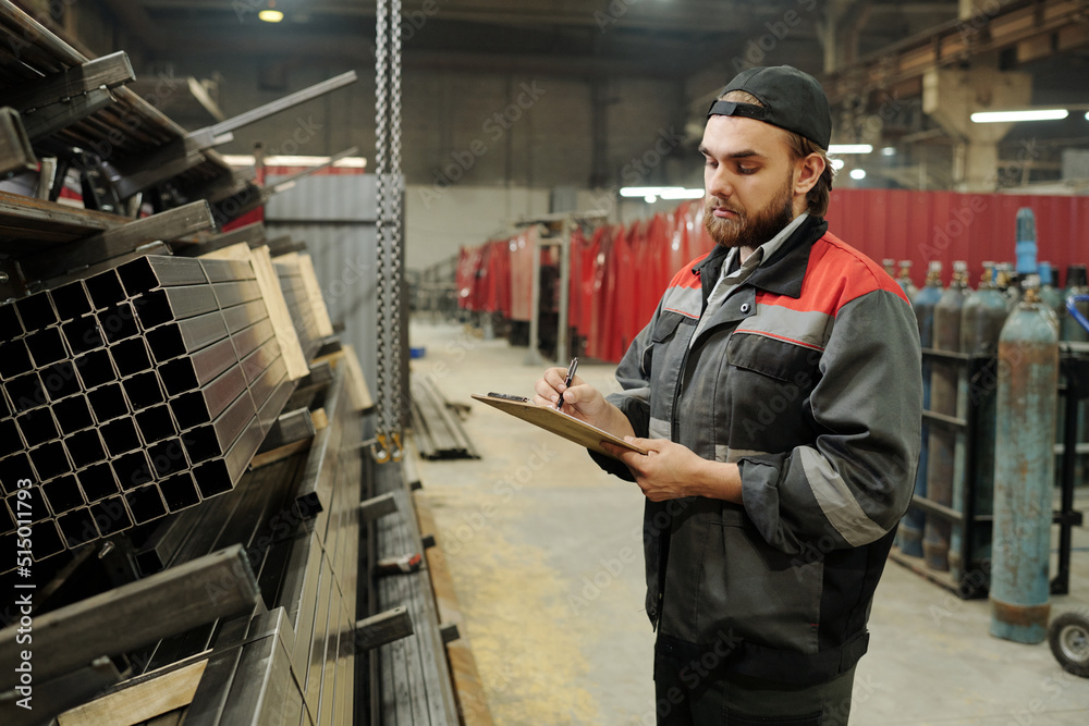 Young male engineer with document making notes about construction materials while standing in front of stack of metallic spare parts