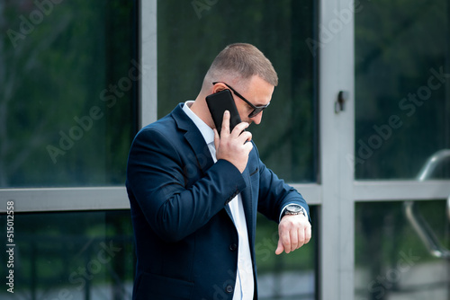 A business man in dark sunglasses with a phone in his hands stands outside the business building talks on the phone and looks at the watch. He is dressed in a light shirt and casual style jacket.