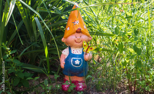 Funny cute colorful garden gnome stands in the garden green grass. Garden dwarf with little butterfly in his hand. Decorative sculpture.