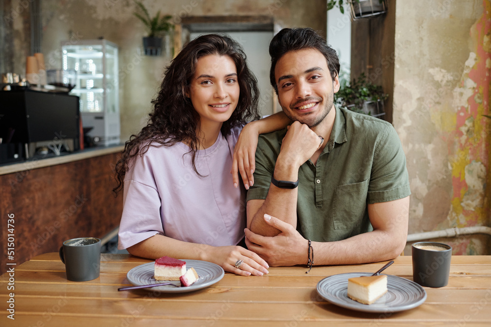 Young cheerful couple looking at camera while sitting by table and having tasty cheesecakes and cappuccino during romantic date in cafe