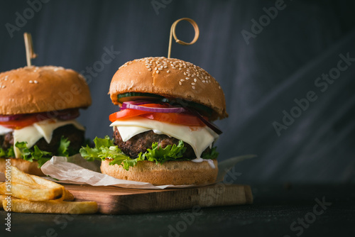 Big beef patty burgers with cheese tomato purple onion cucumber and lettuce next to french fries on wooden black stand on black background