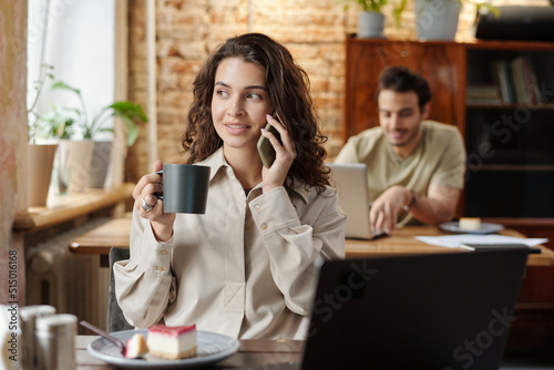 Happy young freelancer talking on mobile phone while sitting by table in front of laptop in cafe and having coffee with cheesecake