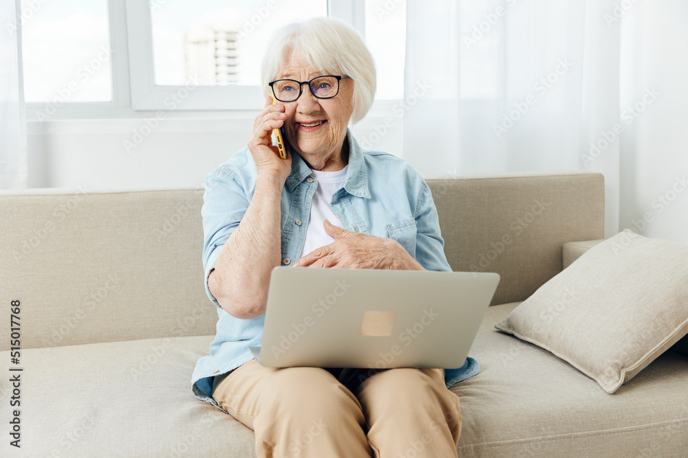 a happy, joyful, pleasant elderly lady is sitting on a cozy sofa in a bright apartment holding a laptop on her lap and happily talking on the phone with her hand leaning against her chest a happy, joyful, pleasant elderly lady is sitting on a cozy sofa in a bright apartment holding a laptop on her lap and happily talking on the phone with her hand leaning against her chest