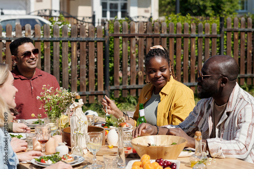 Young black couple and their interracial friends chatting during outdoor dinner by table served with homemade food and drinks
