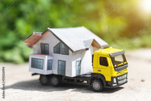 Close-up of a small toy truck on a tree stump carrying a miniature house model on top against a blurred forest background, front view. Countryside relocation service concept. © Angelov