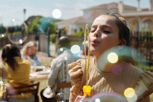 Little girl blowing soap bubbles while playing on backyard of country house against group of intercultural people gathered by table