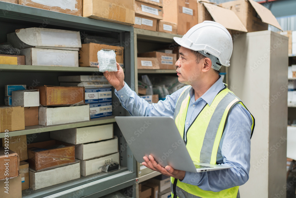 Senior male engineer looking product number of spare part in his hand for checking inventory stock in laptop at warehouse office Senior male engineer looking product number of spare part in his hand for checking inventory stock in laptop at warehouse office