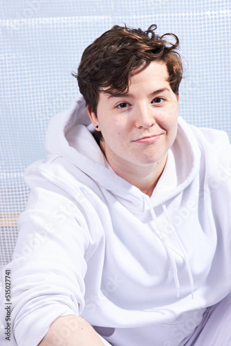 Natural portrait of young handsome transgender man posing with neutral happy face expression. Caucasian with wavy brown hair. Wearing white hoodie and trousers.