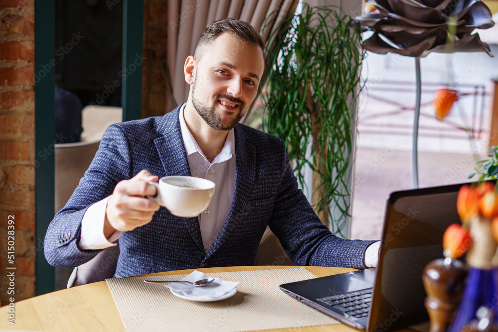 A young man in a business suit sits at a table in a restaurant. Businessman drinking coffee while working at his laptop in a restaurant or cafe A young man in a business suit sits at a table in a restaurant. Businessman drinking coffee while working at his laptop in a restaurant or cafe