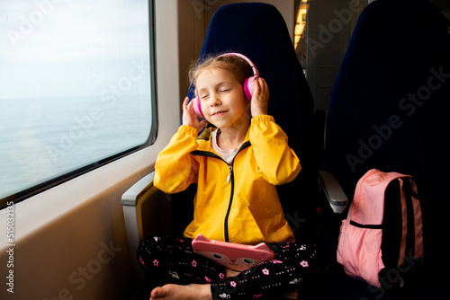 A girl on a train listens to music in headphones with a gadget in her hands. The girl is 6 years old. She goes on vacation with her parents. Family vacation. Gadgets. Vacation. Summer.