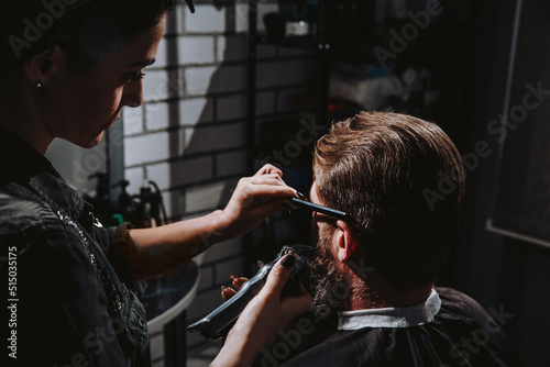 Young woman barber making haircut of bearded man in barbershop. Self-care, masculine beauty