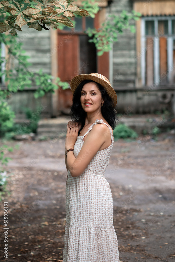 woman in beige dress and straw hat is standing behind the wooden house and smiling woman in beige dress and straw hat is standing behind the wooden house and smiling