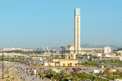 The Great Mosque of Algiers, world's tallest minaret and it's dome seen from Sablettes promenade's ferry wheel. Aerial view of a crowd people walking, blue clear sky and mountains in background.