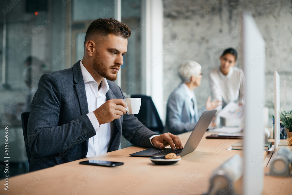 Young businessman drinks coffee while using laptop in office. Young businessman drinks coffee while using laptop in office.