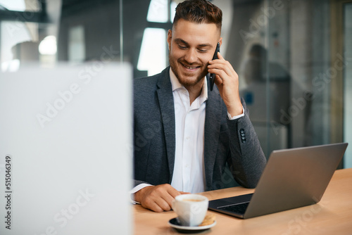 Happy businessman making phone call while working on laptop in office.