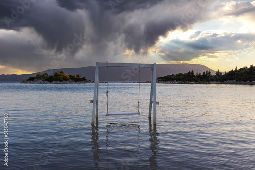 A white wooden swing standing on the edge of a shallow sea, a green island in the background and a dramatic sky with clouds