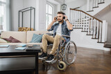 Young man using headphones while sitting in wheelchair at bright living room and listening new songs. Bearded male feeling calmness during relaxation with music at home.