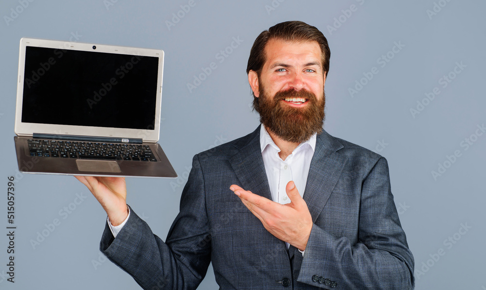 Smiling businessman shows with hand at laptop. Bearded man with computer. Blank screen on notebook. Smiling businessman shows with hand at laptop. Bearded man with computer. Blank screen on notebook.