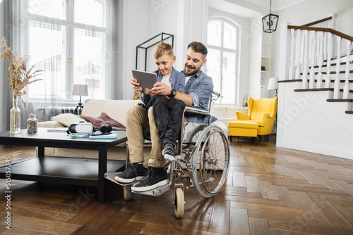 Smiling dad with special needs sitting in wheelchair and holding son on knees while playing on digital tablet. Positive family using portable device for playing childrens educational games.