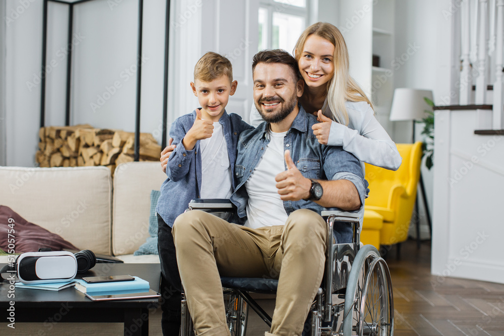 Smiling disabled man in wheelchair posing on camera together with cute son and pretty happy wife in apartment with modern interior. Concept of happy family and support. Smiling disabled man in wheelchair posing on camera together with cute son and pretty happy wife in apartment with modern interior. Concept of happy family and support.