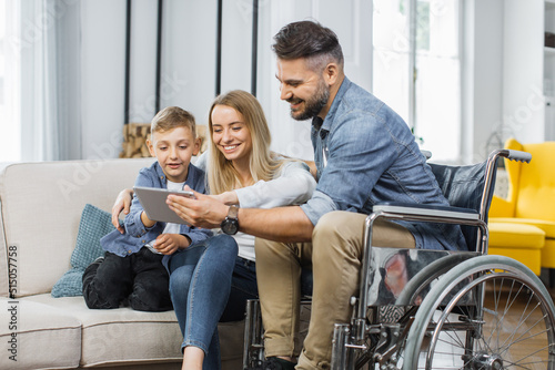 Caucasian man in wheelchair and his lovely wife with cute son watching video on digital tablet at home. Concept of family, disability and technology.
