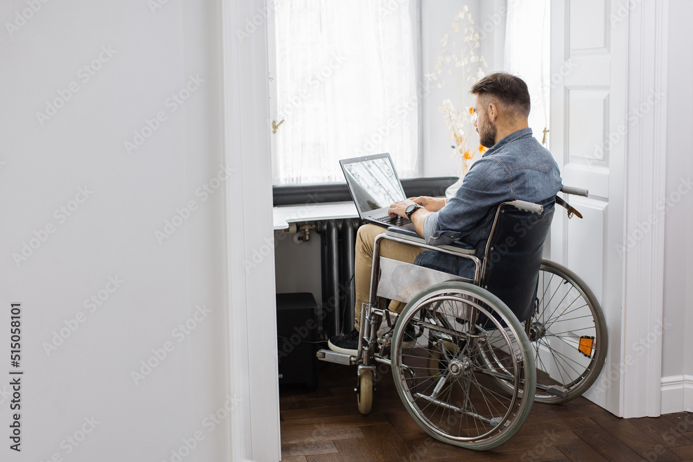 Confident bearded man who using wheelchair typing on wireless laptop at home. Disabled freelancer in casual wear working and chatting remotely with clients. Confident bearded man who using wheelchair typing on wireless laptop at home. Disabled freelancer in casual wear working and chatting remotely with clients.