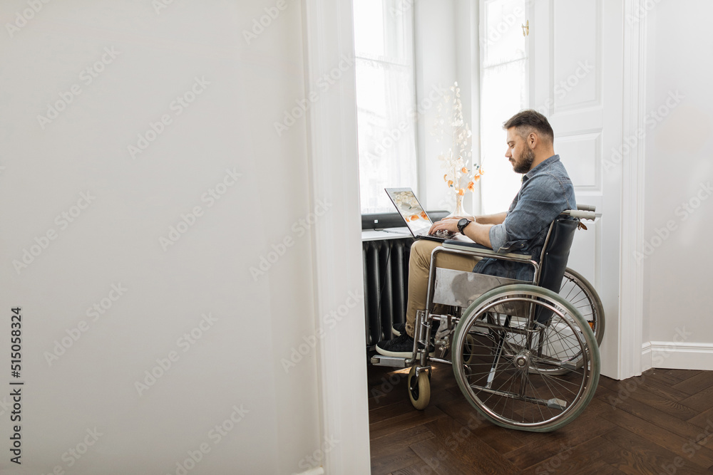 Serious disabled young man using laptop for remote job at home. Confident caucasian male in wheelchair surfing internet near window at workplace. Serious disabled young man using laptop for remote job at home. Confident caucasian male in wheelchair surfing internet near window at workplace.