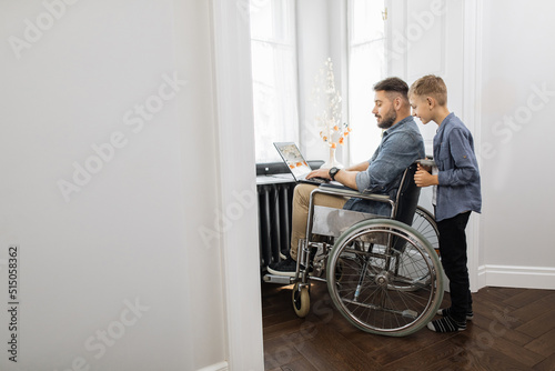 Lovely son standing behind dad that sitting in wheelchair with modern laptop on knees. Caucasian bearded man and school age boy spending time together at home.